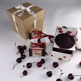 Angelic Thoughts Brown Kraft Gift Box Tied with Angelic Thoughts Ribbon,Cherish Love Cherry Love Gift Box Bundle Standard being displayed with loose cherries scattered amongst products which are: 2 x Cherry Soft Nougat, 2 x Cherry & Macadamia Nougat, Hand poured Soy Wax Cherry Candle in Art Deco Jar with lid on