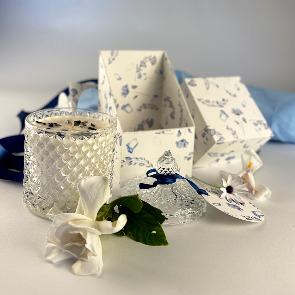 Floral handpoured soy wax candle in Art Deco Crystal Glass embellished with lapus lazuli. The lid iwith a label tied in navy blue ribbons sitting beside the candle infront of an open product box with a rustal and floral print and fresh flowers are scattered