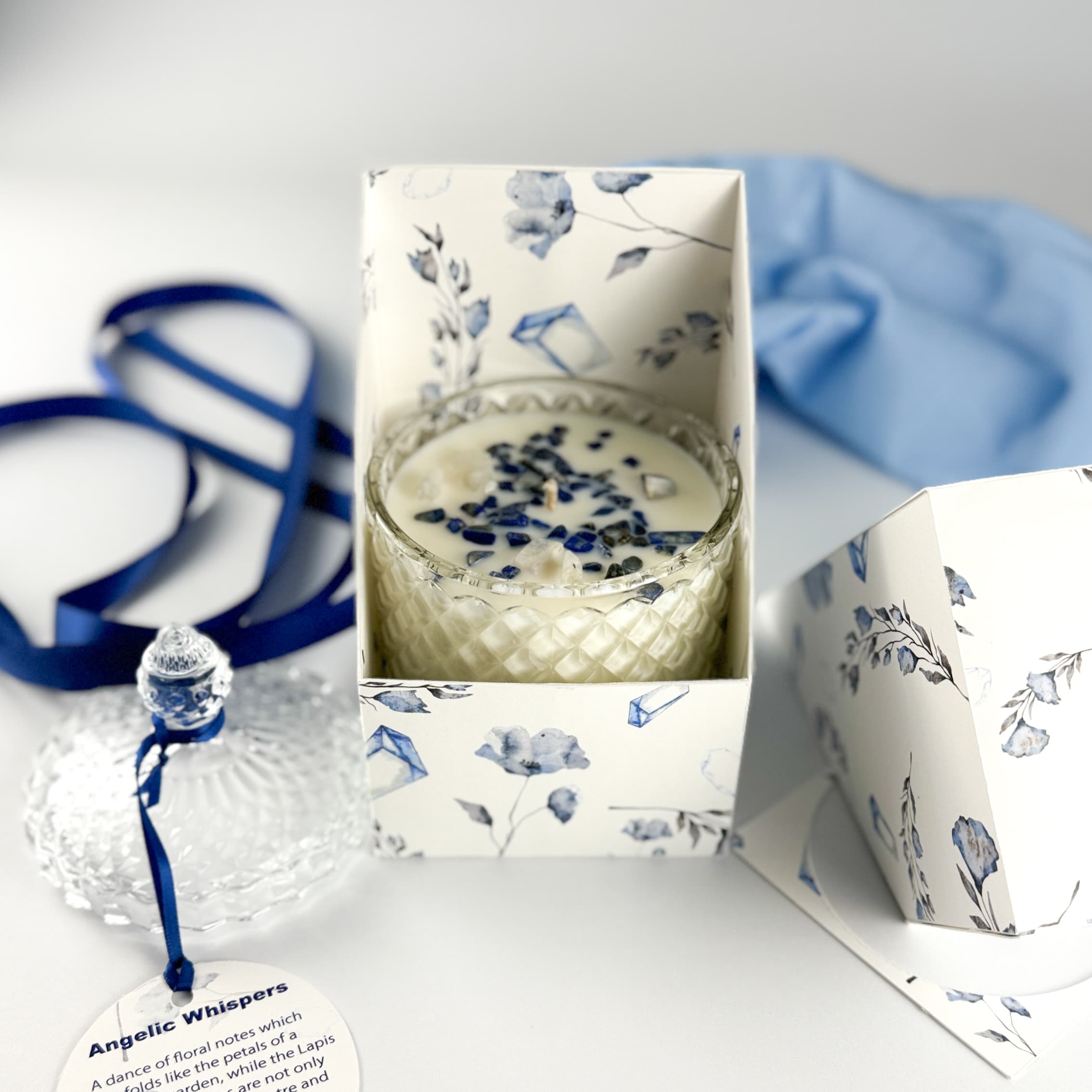 Angelic Whispers floral handpoured soy wax candle in Art Deco Crystal Glass embellished with lapus lazuli. The lid is off and beside it with a label tied in navy blue ribbons sitting beside the candle infront of an open product box with a rustal and floral print