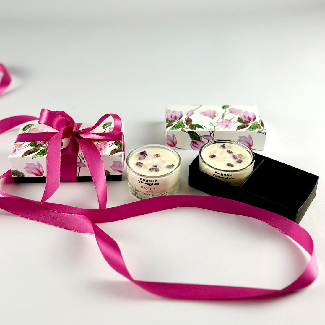 magnolia scented rose quartz and amethyst embellished hand poured tealights in glass tealight containers - one in black base box and one out, with product lid behine and with magnolia 2pack tealight product box tied with deep pink ribbon on the left side