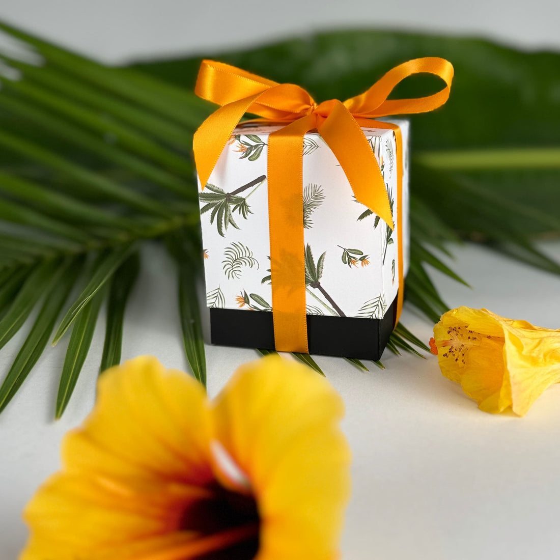 Golden orange hibiscus in foreground with Paradise Whispers product box featuring palm trees and bird of paradise flowers and black base tied in orange ribbon