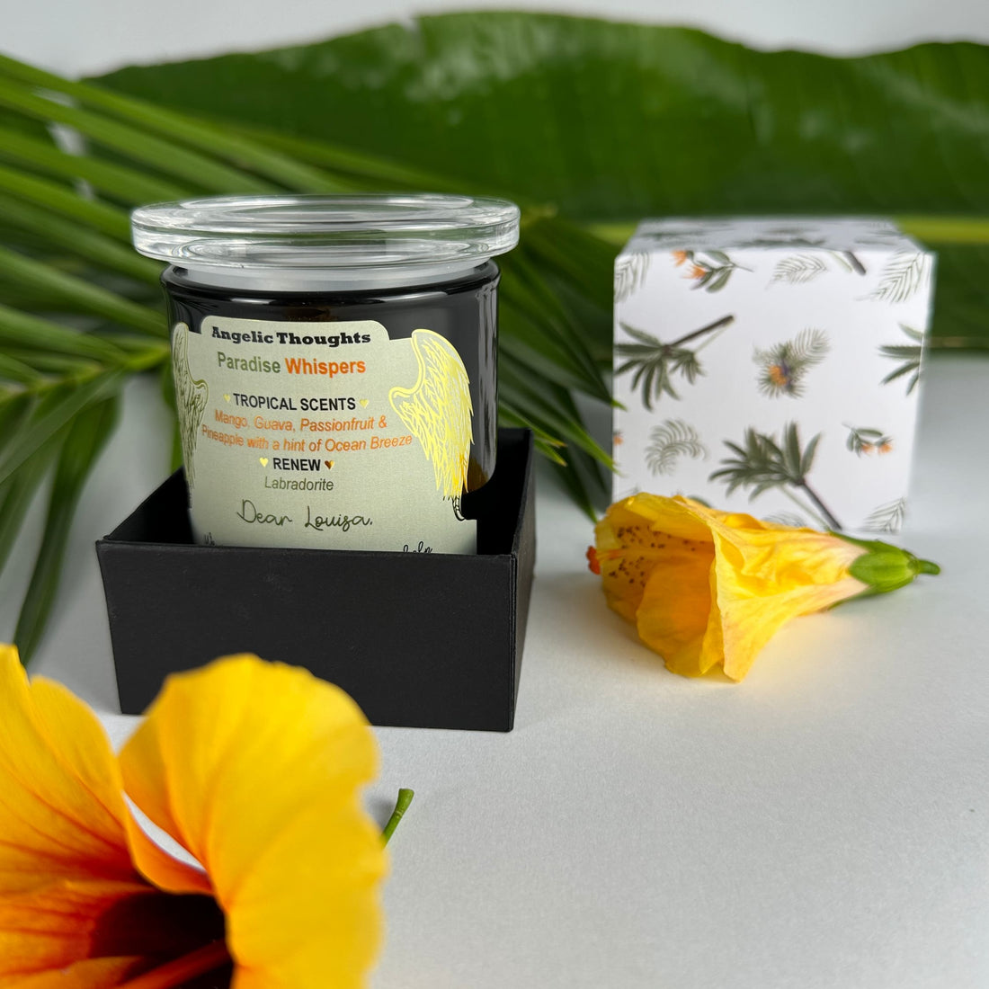 Golden orange hibiscus in foreground with Paradise Whispers candle in box base and lid featuring palm trees and bird of paradise flowers and closed hibiscus flower in background 