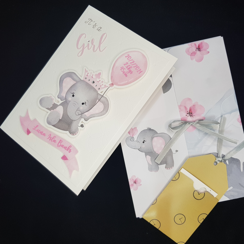 Image of the baby girl grey elephant card and a folder. The image of baby girl card is on the left. The Folder image has an image of a small  grey elephant, scattered pink flowers and a grey ribbon tie to keep folder closed. There is also an image of the gift tag holder containg the white gift tag. The holder is a mustard colour.