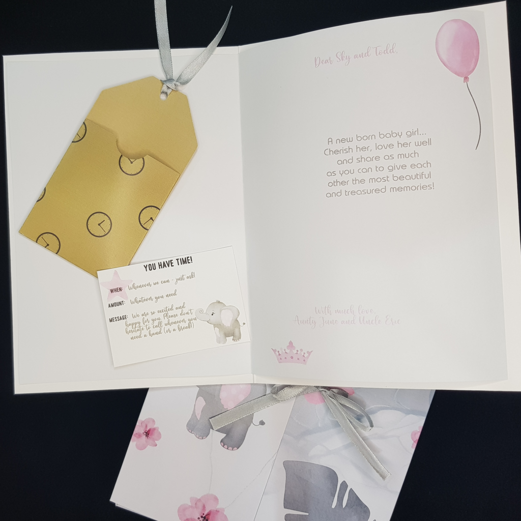 An Image of the open card with a Gift Tag holder including an example gift tag is on the left hand side of image. The image of the gift tag has a small grey elephant on it. A lovely verse is on the right hand side of the image, as well as a pink balloon image in the top right corner. In the bottom left corner is a small pink crown. The matching folder is partially hidden behind the card and has images of baby elephant and pink flowers on it. It is tied with a grey ribbon.
