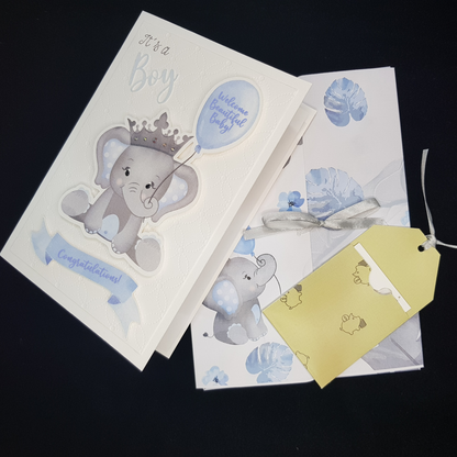 Image of the baby boy grey elephant card and a folder. The image of baby boy card is on the left. The Folder image has an image of a small grey elephant, scattered blue and grey leaves and a grey ribbon tie to keep folder closed. There is also an image of the gift tag holder containing the white gift tag. The holder is a mustard colour.