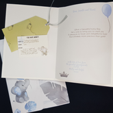An Image of the open card with a Gift Tag holder including an example gift tag is on the left hand side of image. The image of the gift tag has a small grey elephant on it. A lovely verse is on the right hand side of the image, as well as a blue balloon image in the top right corner. In the bottom left corner is a small silver crown. The matching folder is partially hidden behind the card and has images of baby elephant and blue and grey leaves on it. It is tied with a grey ribbon.