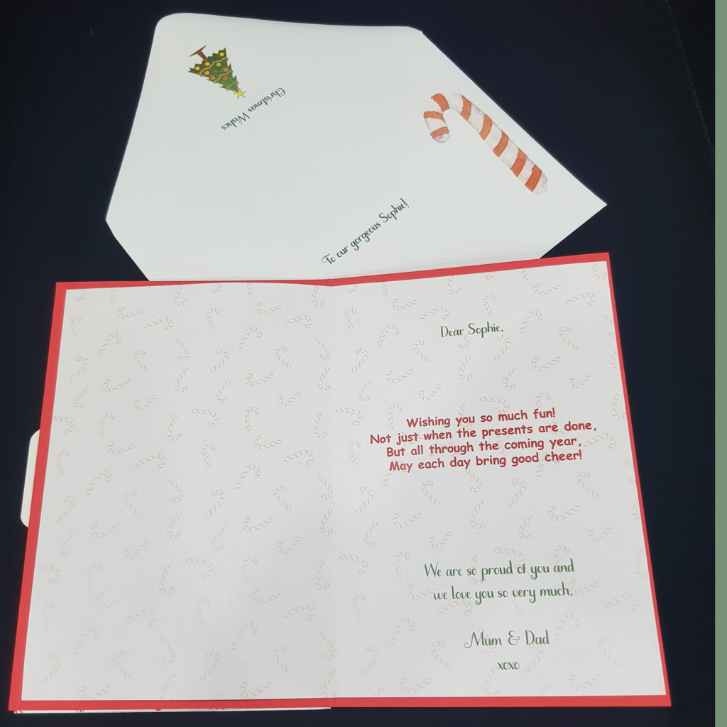 Opened Christmas Greeting card. Red card stock with white paper insert. Text shown. Envelope behind with Candy cane and Christmas tree.