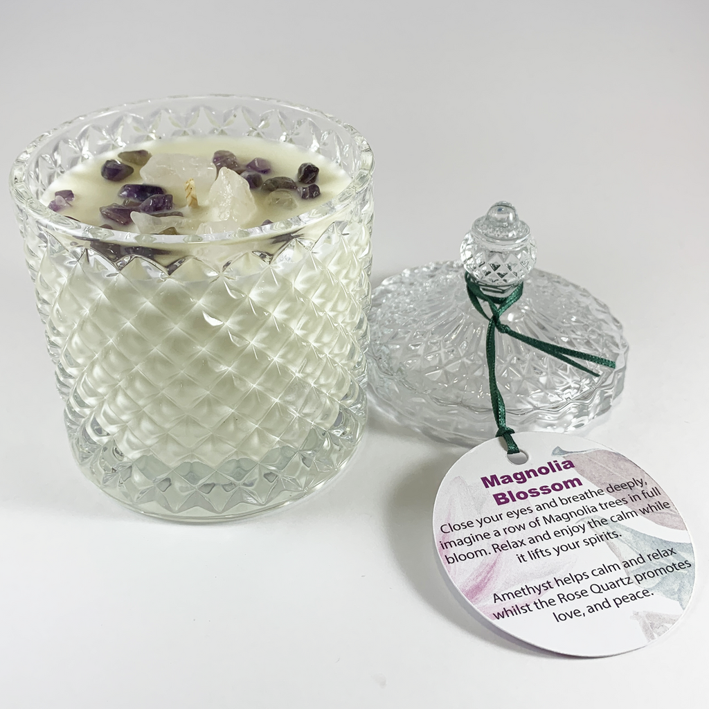 magnolia scented rose quartz and amethyst embellished hand poured soy wax candle in clear glass art deco style container with lid off to the right. Magnolia Blossom product label is shown face up on the bottom right