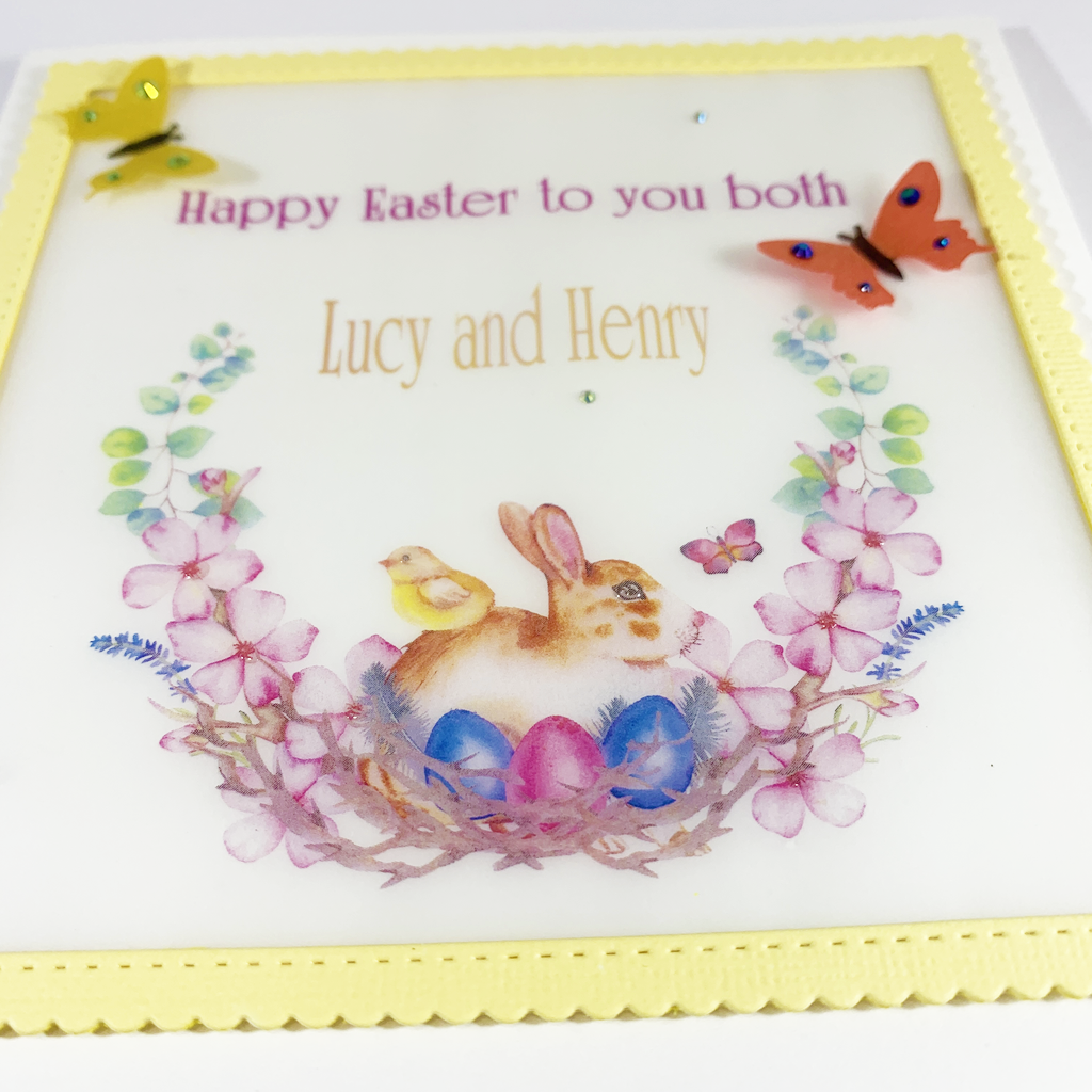 This image is a close up of the front of the card at an angle allowing you to see the elevation of the butterflies. You can also see the detail in the nest and flowers the bunny is sitting in. The words on the front of the card read &quot;Happy Easter to you both&quot; on one line and on another line &quot;Lucy and Henry&quot;