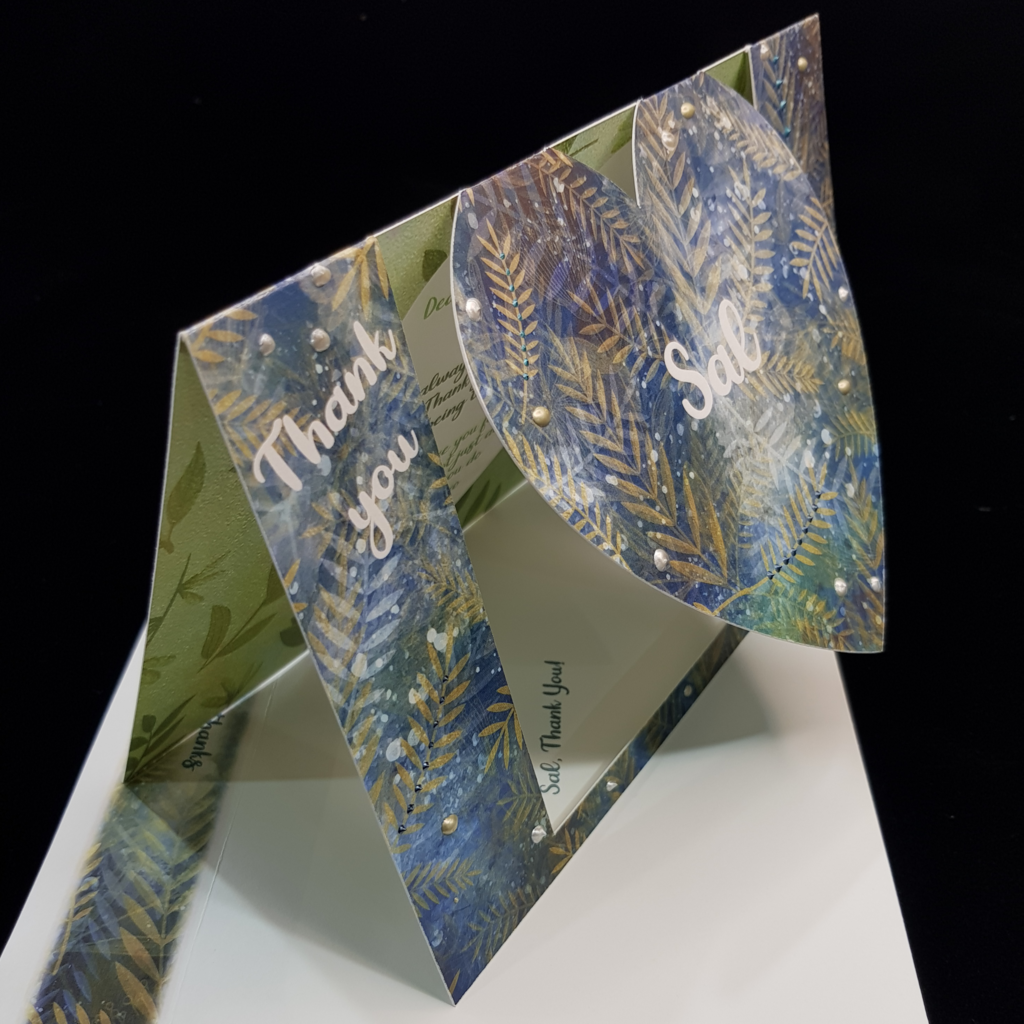 A beautiful heart swing card stands upright on a matching envelope. with the words &quot;thank you&quot; on the left, the name Sal sits centrally on the swinging heart and another sentiment &quot;so very much&quot; sits on the right. Delicate golden stems of foliage overlay over the top of a mottled blue toned background. Pops of pearl colours ranging from white, antique gold and blues have been selectively hand applied.