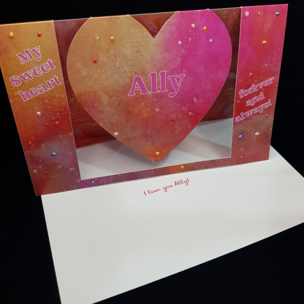 A beautiful heart swing card stands upright on a matching envelope with the words &quot;I love you Ally! on the front. The card features the words &quot;My Sweet heart&quot; on the left, the name Ally sits centrally on the swinging heart and another sentiment &quot;forever and always&quot; sits on the right. a splotchy mix of bright pink and sherbert orange forms the focus of this bright mixed tone background. Pops of clear and pearl colours including orange, purple red and pink have been selectively hand applied.