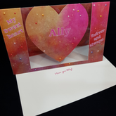 A beautiful heart swing card stands upright on a matching envelope with the words "I love you Ally! on the front. The card features the words "My Sweet heart" on the left, the name Ally sits centrally on the swinging heart and another sentiment "forever and always" sits on the right. a splotchy mix of bright pink and sherbert orange forms the focus of this bright mixed tone background. Pops of clear and pearl colours including orange, purple red and pink have been selectively hand applied.