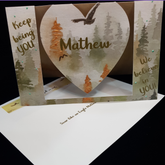 A beautiful heart swing card sits open on a matching envelope.  The words " Keep being you" on the left, the name Mathew sits centrally on the swinging heart and another sentiment "We believe in you" sits on the right. This card features a misty watercolor pine forest scene in muted autumn tones with soaring birds.  The main bird has been embellished with clear accents and the orange trees have been embellished with an auburn pearl colour. There are spots of page green dotted on the card for interest.