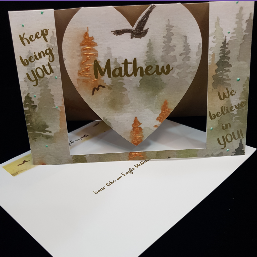 A beautiful heart swing card sits open on a matching envelope.  The words &quot; Keep being you&quot; on the left, the name Mathew sits centrally on the swinging heart and another sentiment &quot;We believe in you&quot; sits on the right. This card features a misty watercolor pine forest scene in muted autumn tones with soaring birds.  The main bird has been embellished with clear accents and the orange trees have been embellished with an auburn pearl colour. There are spots of page green dotted on the card for interest.