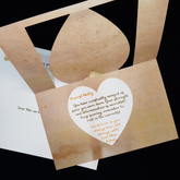 An open heart swing card sits on a matching envelope. A white heart slightly smaller than the cutout heart sits centrally overlaid on a background of beige to antique gold splotched graduated background. A white heart slightly smaller that the cutout heart sits centrally overlaid on the background. The name and sign off is written in an orange colour with the verse in a deep green. Fully customisable