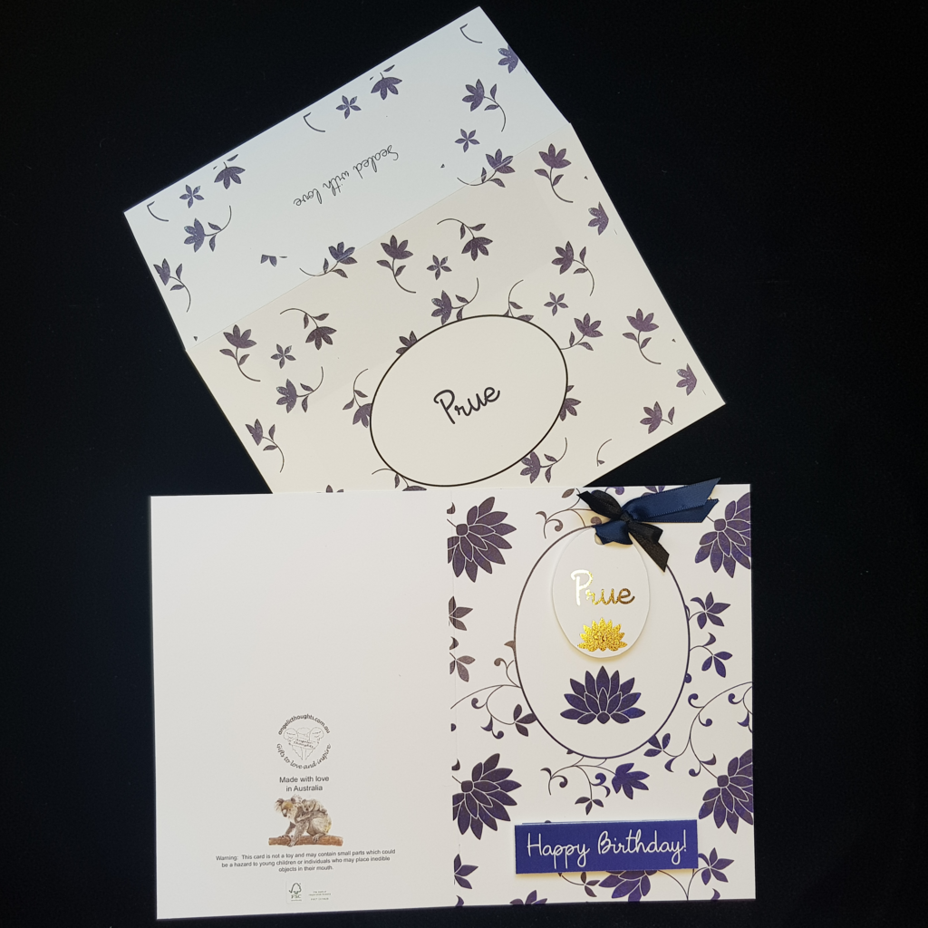The card is open flat with the outside showing. On the left is the back of the card featuring the Angelic Thoughts Logo, and a Choking Hazard Warning, on the right is the front of the card - refer to Front of card alt text.  The card is laying on an open envelope. Stylised blue flowers cover the envelope. A  blue oval ring sits in the centre to hold the recipients name / brief message. On the back of envelope a plain white oval holds a brief message